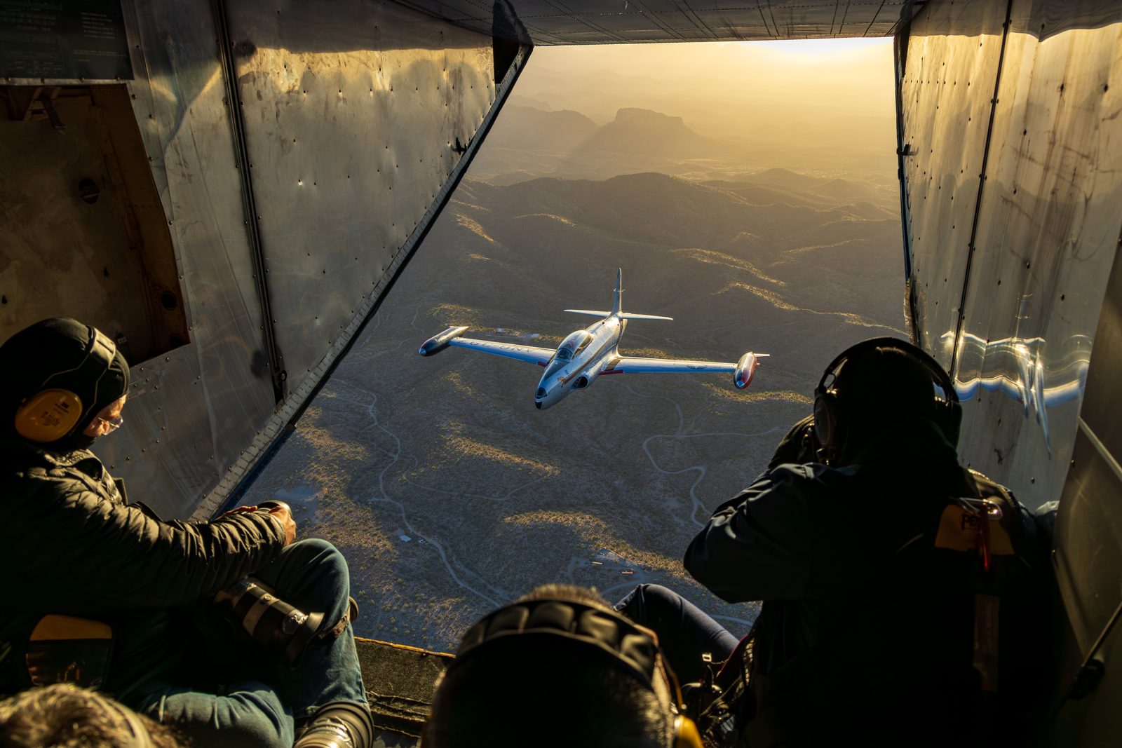 Two photographers with long lenses lean into the open door of a camera aircraft, a jet visible in the distance beyond.