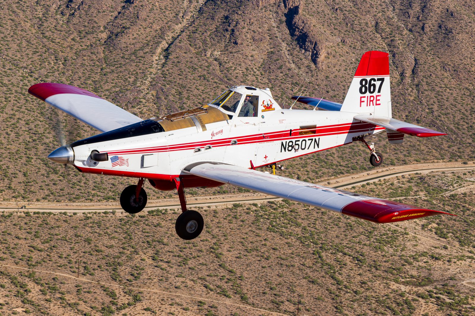 Red and white Air Tractor AT-802A firefighting aircraft marked 867 FIRE, banking over rocky desert.